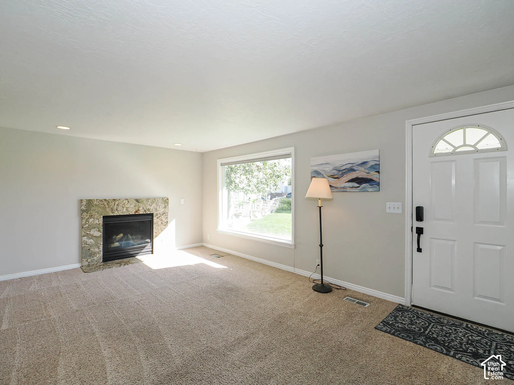 Neutral tone wall paint, carpet flooring, stone-surround fireplace, large window with outdoor visibility, and a white front door with an arched transom