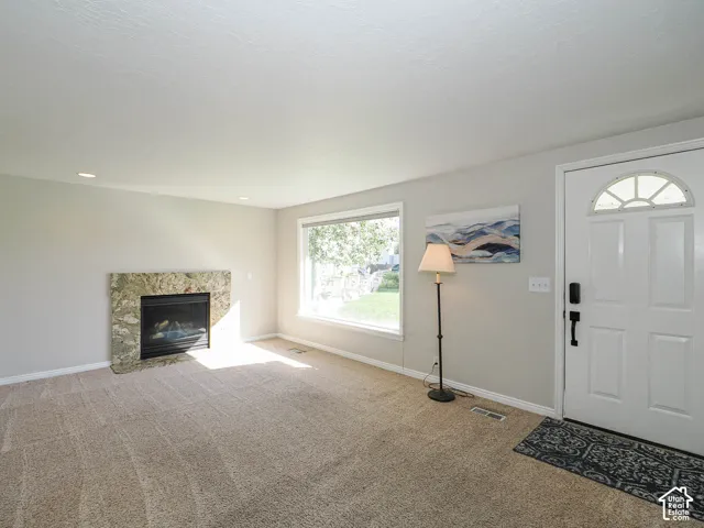 Neutral tone wall paint, carpet flooring, stone-surround fireplace, large window with outdoor visibility, and a white front door with an arched transom