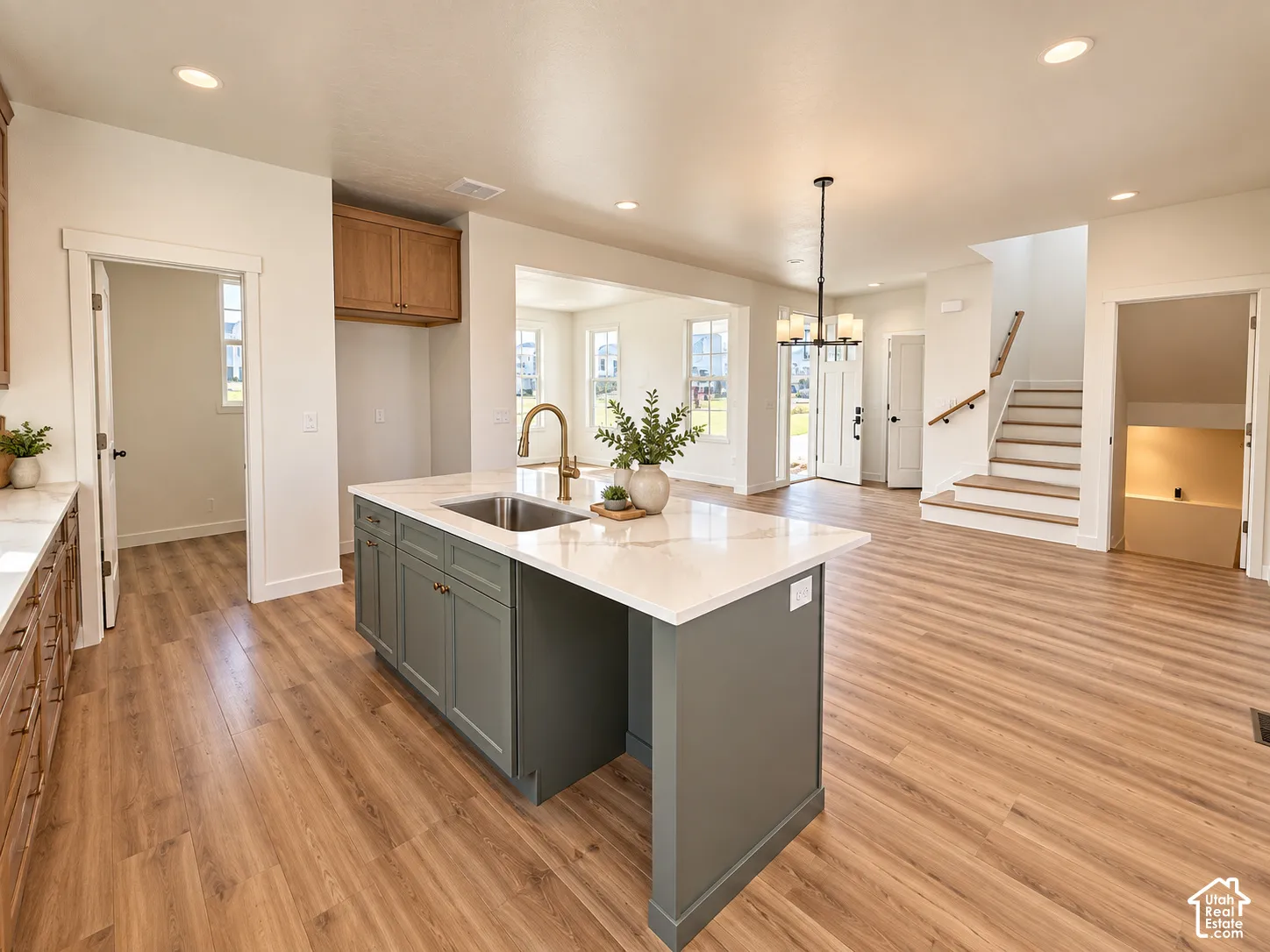Kitchen island featuring a single-basin sink and brass faucet, white countertop, wood-finish flooring, recessed lighting, and a staircase with wood handrail