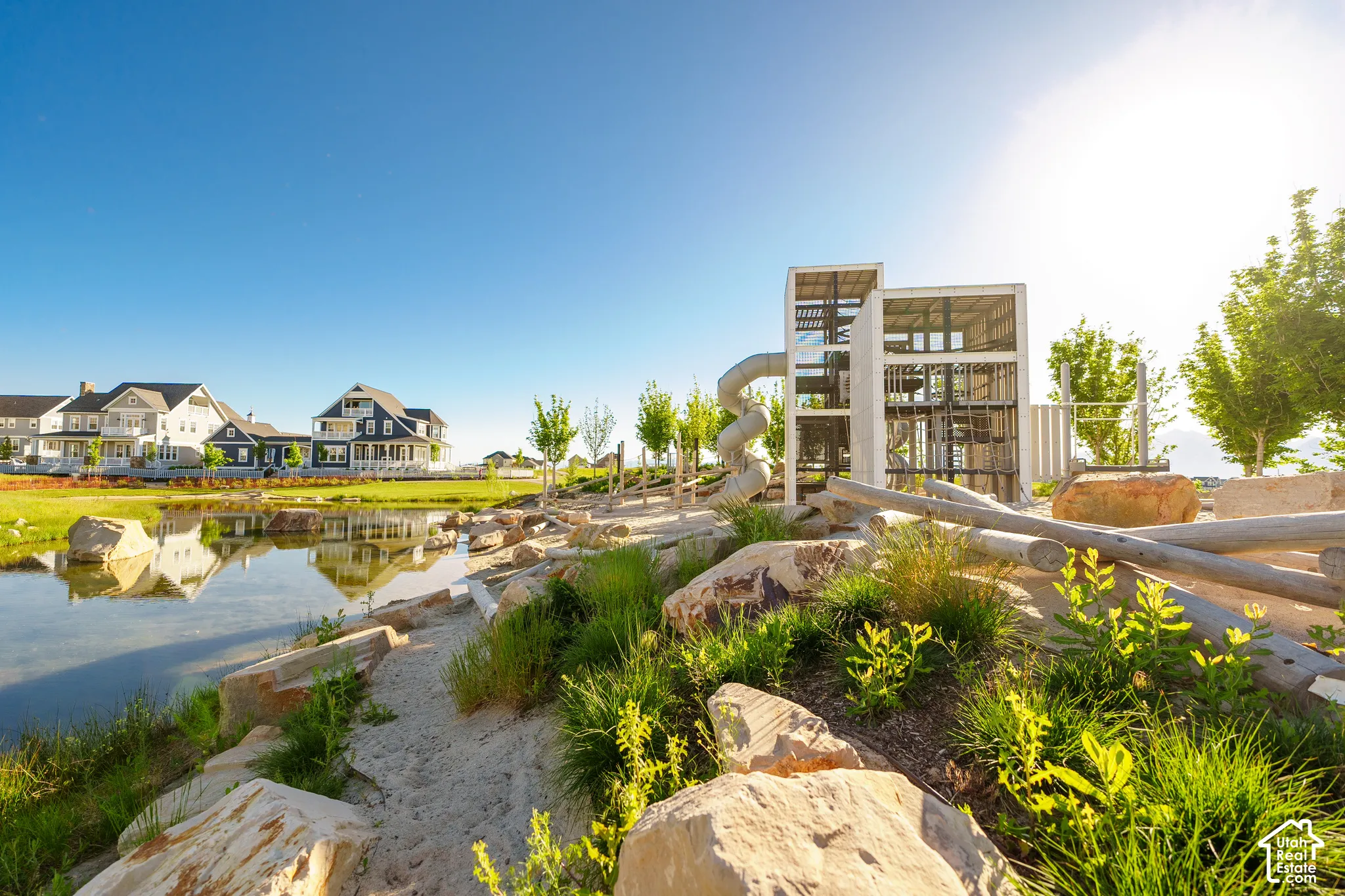 Community pond with surrounding green space, a contemporary play structure, natural rock landscaping, and distant residential properties