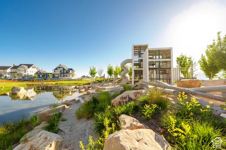 Community pond with surrounding green space, a contemporary play structure, natural rock landscaping, and distant residential properties