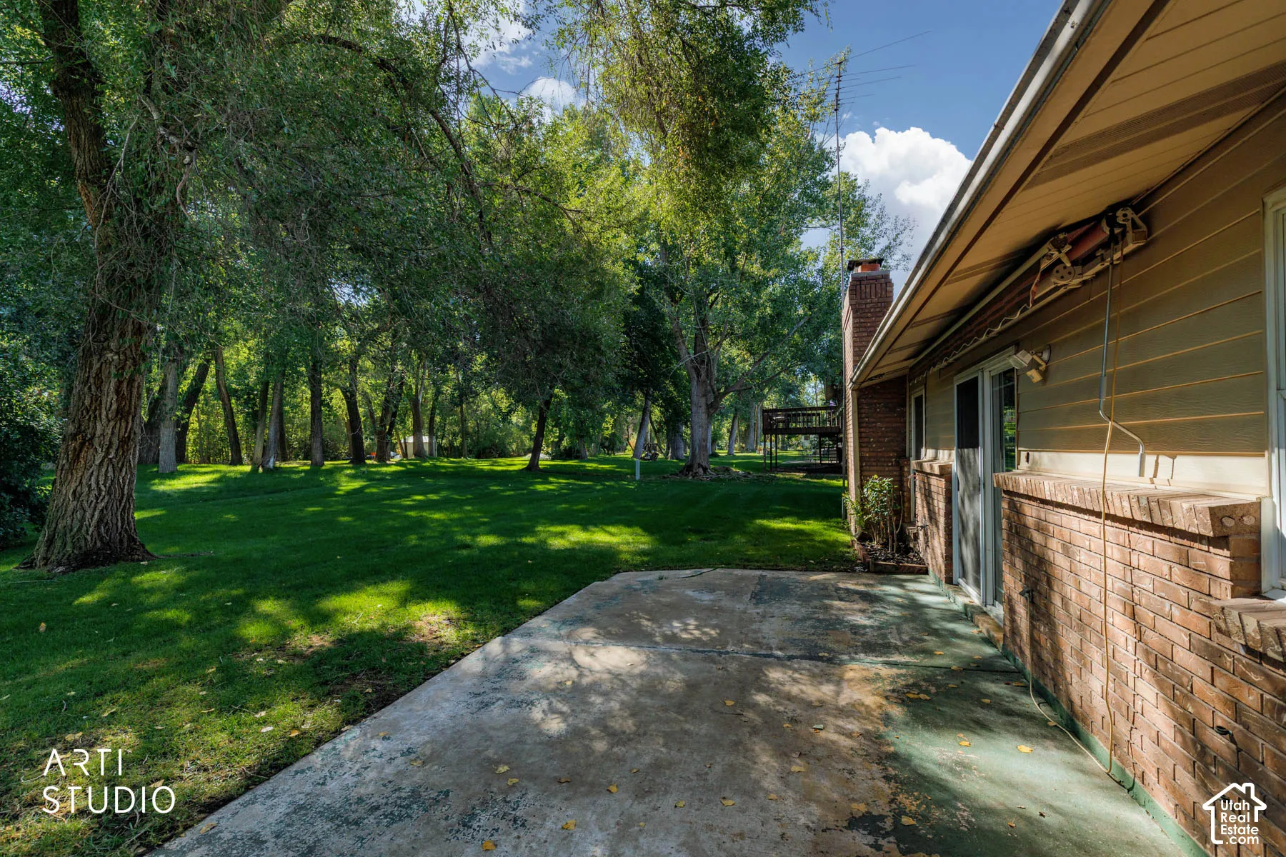 Expansive green space featuring mature trees, a concrete patio, brick exterior, and a sliding glass door