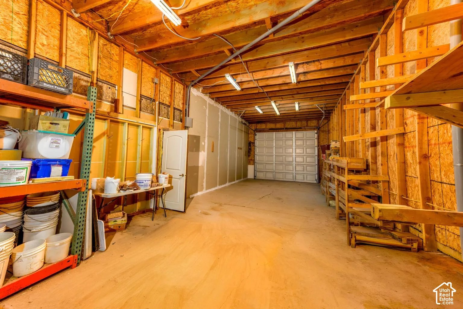Spacious garage interior featuring exposed wood framing, a white overhead garage door, built-in shelving, a side door, and fluorescent lighting
