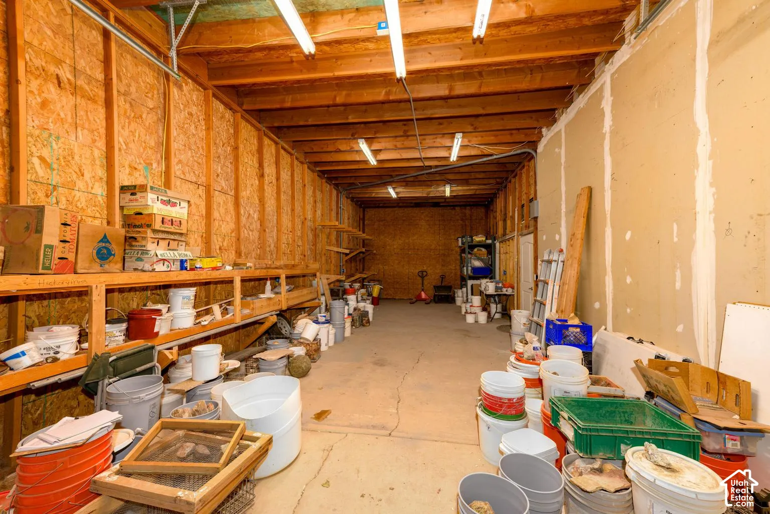 Exposed wood beam ceiling, fluorescent lighting fixtures, concrete flooring, unfinished plywood walls, and unfinished drywall walls