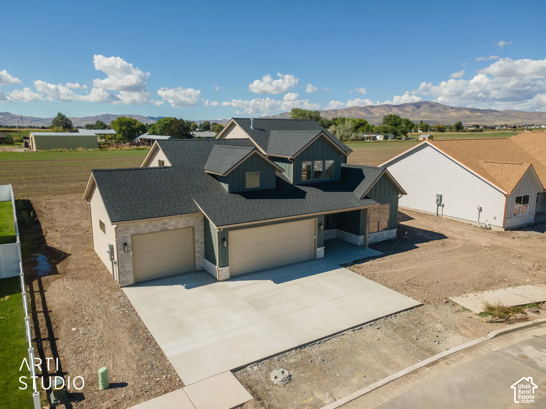 Multi-pitch dark shingle roof, two-car garage with light tan doors, stone facade accents, dark vertical siding, and a concrete driveway