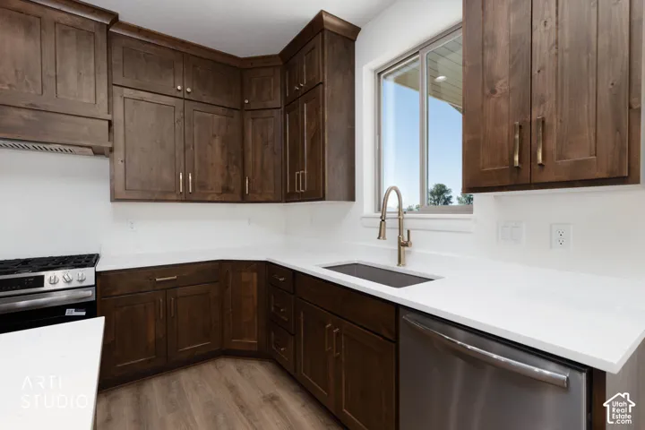 Dark wood-finish cabinetry, white solid surface countertops, stainless steel appliances, wood-finish flooring, and a stainless steel sink with a brushed gold faucet