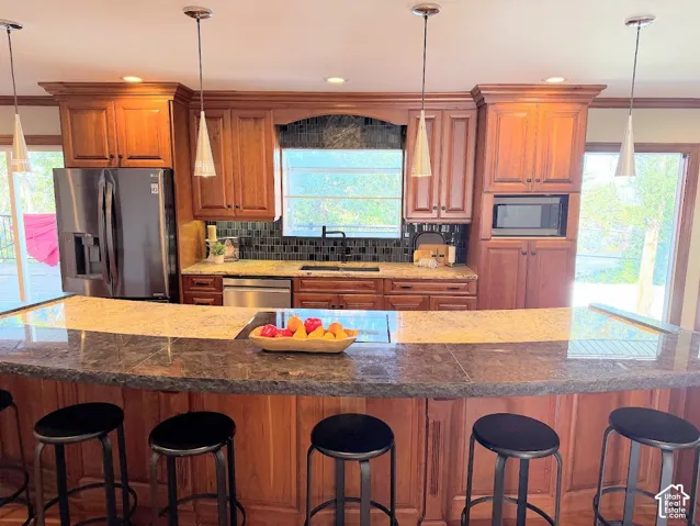Kitchen featuring wood cabinetry, stainless steel appliances, a dark tile backsplash, and a multi-level island with a stone countertop