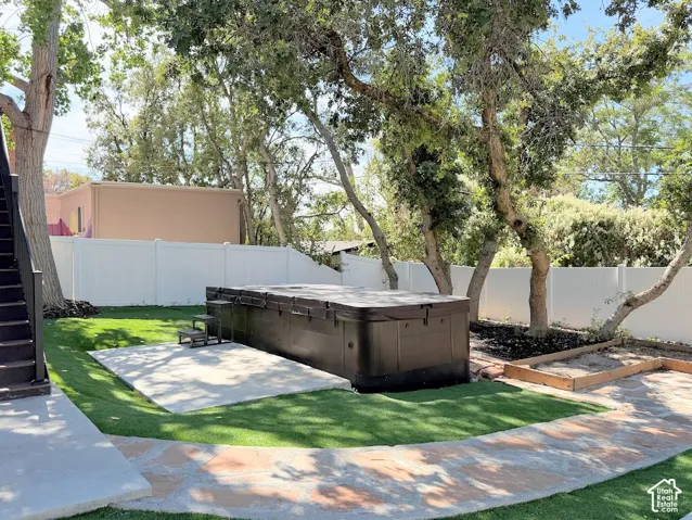 Lush artificial turf surrounds a concrete patio, a large spa, and a raised garden bed, all enclosed by a white privacy fence