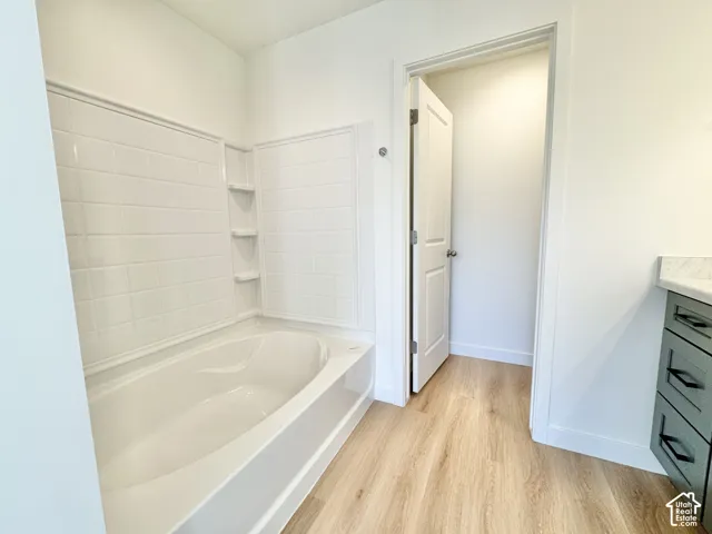 Bathtub with tiled surround and built-in shelving, wood-finish flooring, and a vanity with a white countertop and dark cabinetry