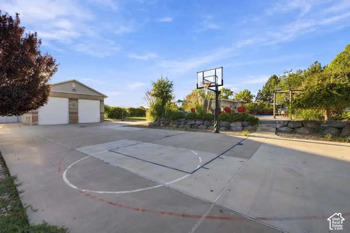 Expansive concrete pad featuring a basketball hoop and court markings, accompanied by a two-car garage with white garage doors and brick accents, and a built-in stone retaining wall