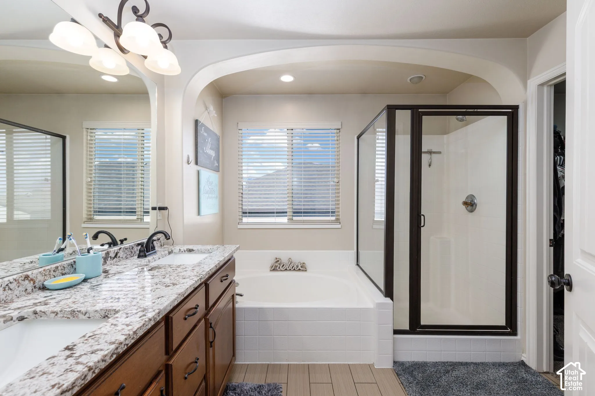 Double vanity with granite-style countertop, wood-finish cabinetry, built-in bathtub with tiled surround, and separate glass-enclosed shower