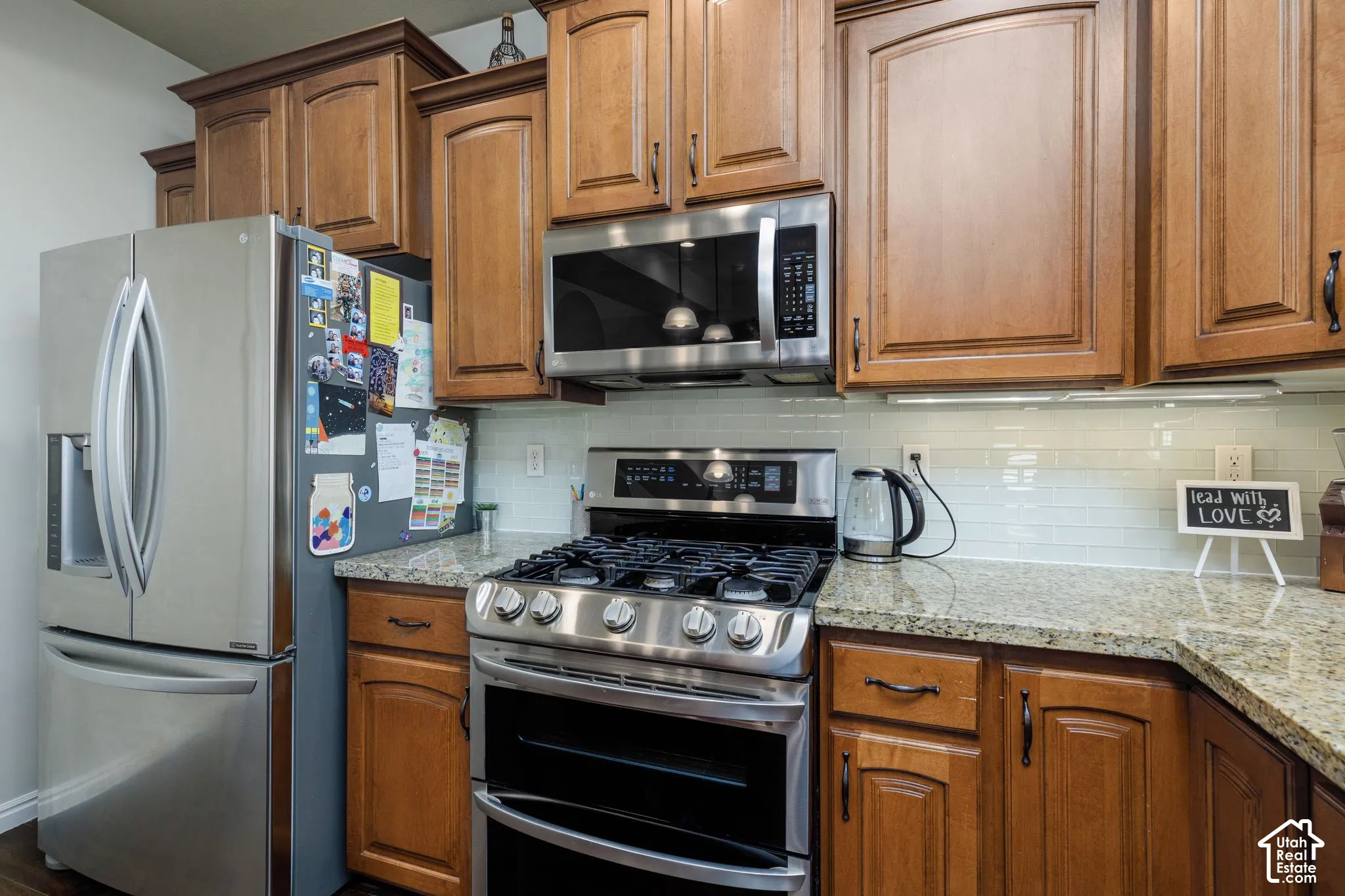 Wood-finish cabinetry, stainless steel range with gas cooktop, stainless steel microwave, light-colored granite countertops, and a white subway tile backsplash