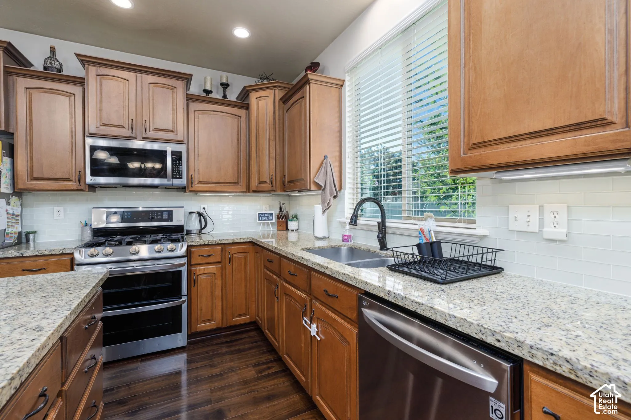 Kitchen featuring wood-finish cabinetry, light-toned granite countertops, stainless steel appliances, a subway tile backsplash, and wood-finish flooring