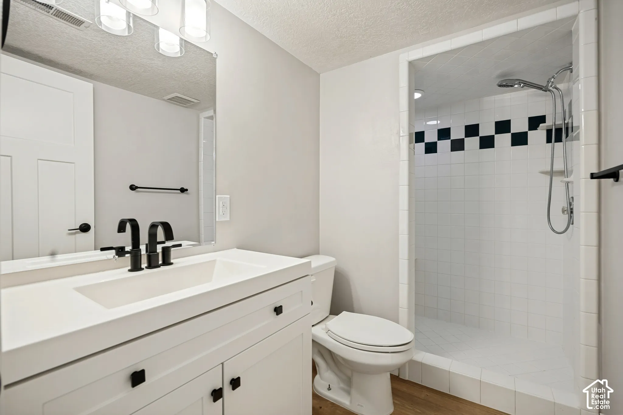 Walk-in shower with checkerboard accent tile, white vanity with integrated sink, black dual-handle faucet, and wood-finish flooring