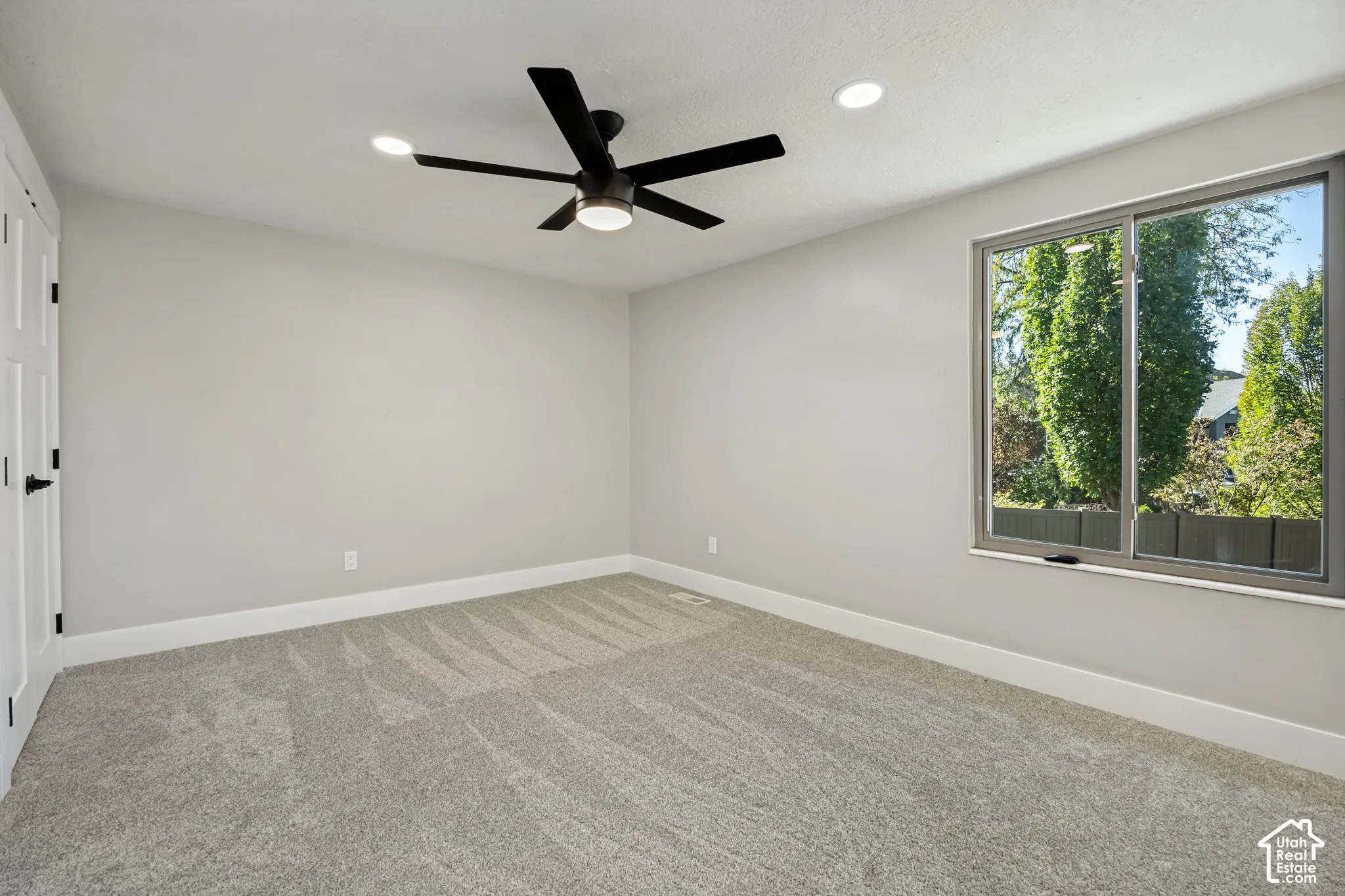 Neutral wall color, gray carpeting, modern ceiling fan, recessed lighting, and a large window with exterior landscaping