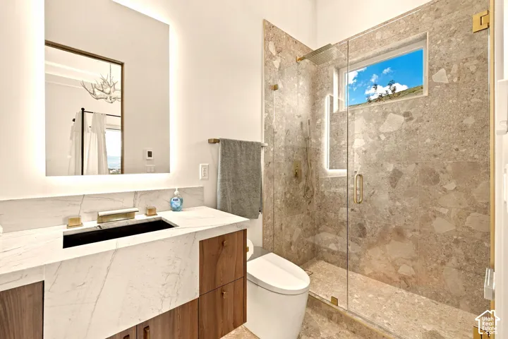 Vanity with integrated sink and wood-finish cabinetry, illuminated mirror, walk-in shower with glass enclosure, built-in shower window, and floor-to-ceiling stone-look wall tiling