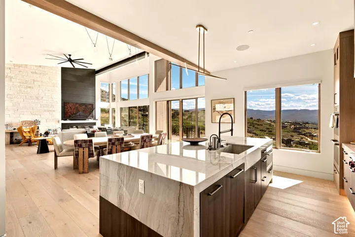 Spacious open-concept living area featuring high ceilings, wood-finish flooring, a stone accent wall with a fireplace, a kitchen island with an undermount sink, and extensive glazing