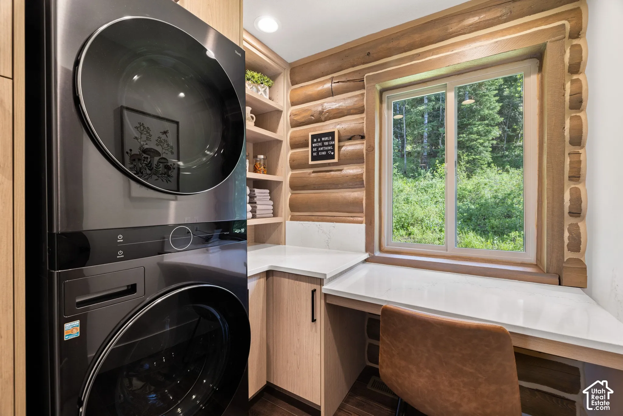 Built-in stacked laundry appliances, light wood-finish cabinetry, white countertop, and log wall accents