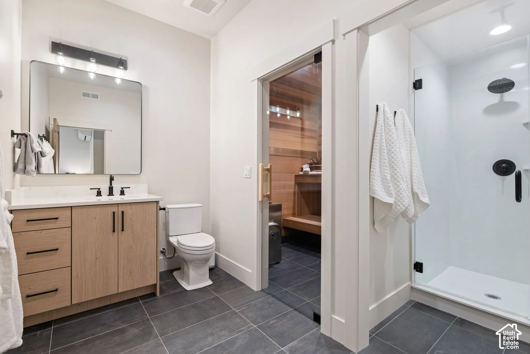 Modern bathroom featuring a wood-finish vanity with a light-toned countertop, a frameless shower with a glass enclosure, and dark tile flooring