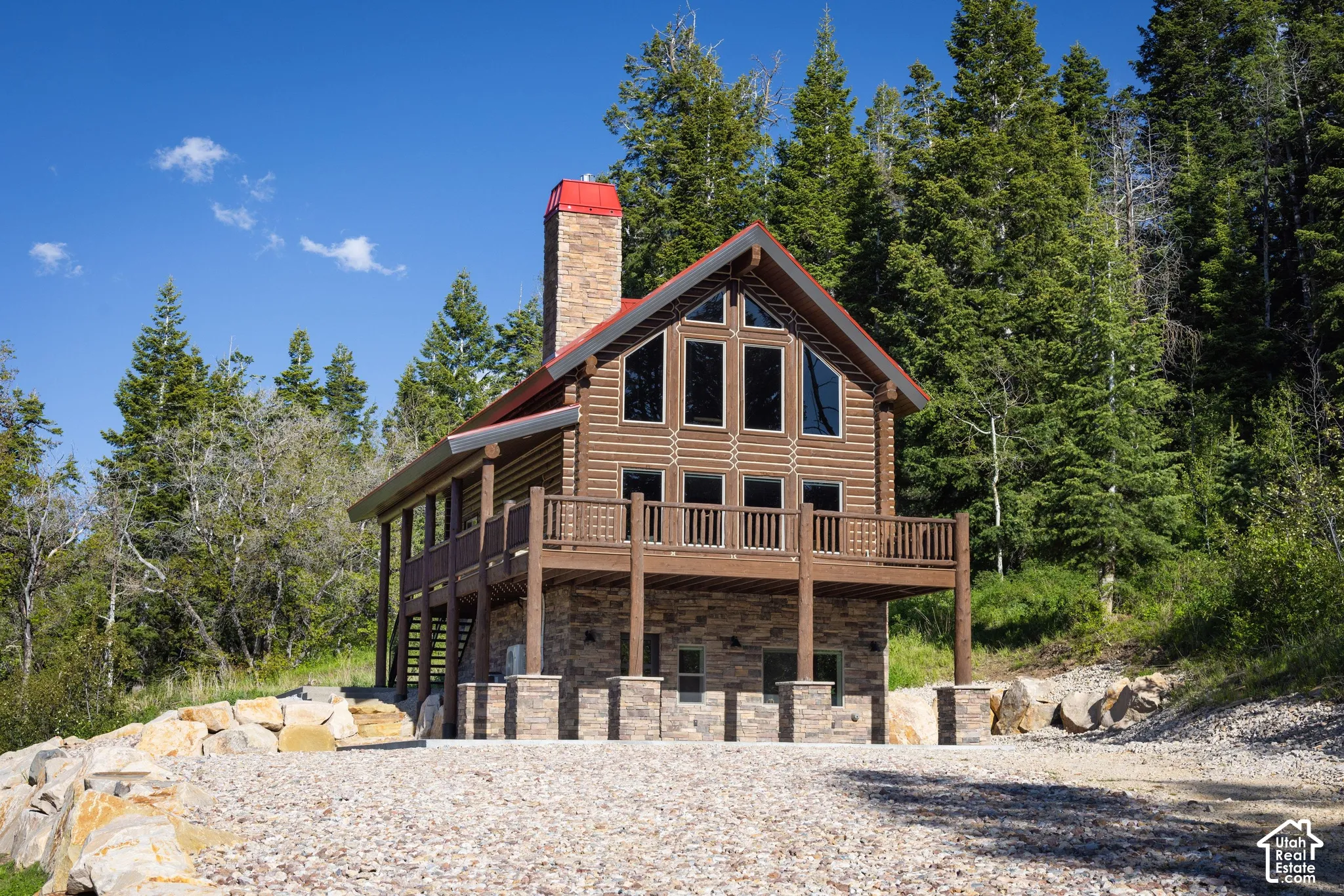 Log cabin exterior with an elevated deck, stone chimney, and gable windows
