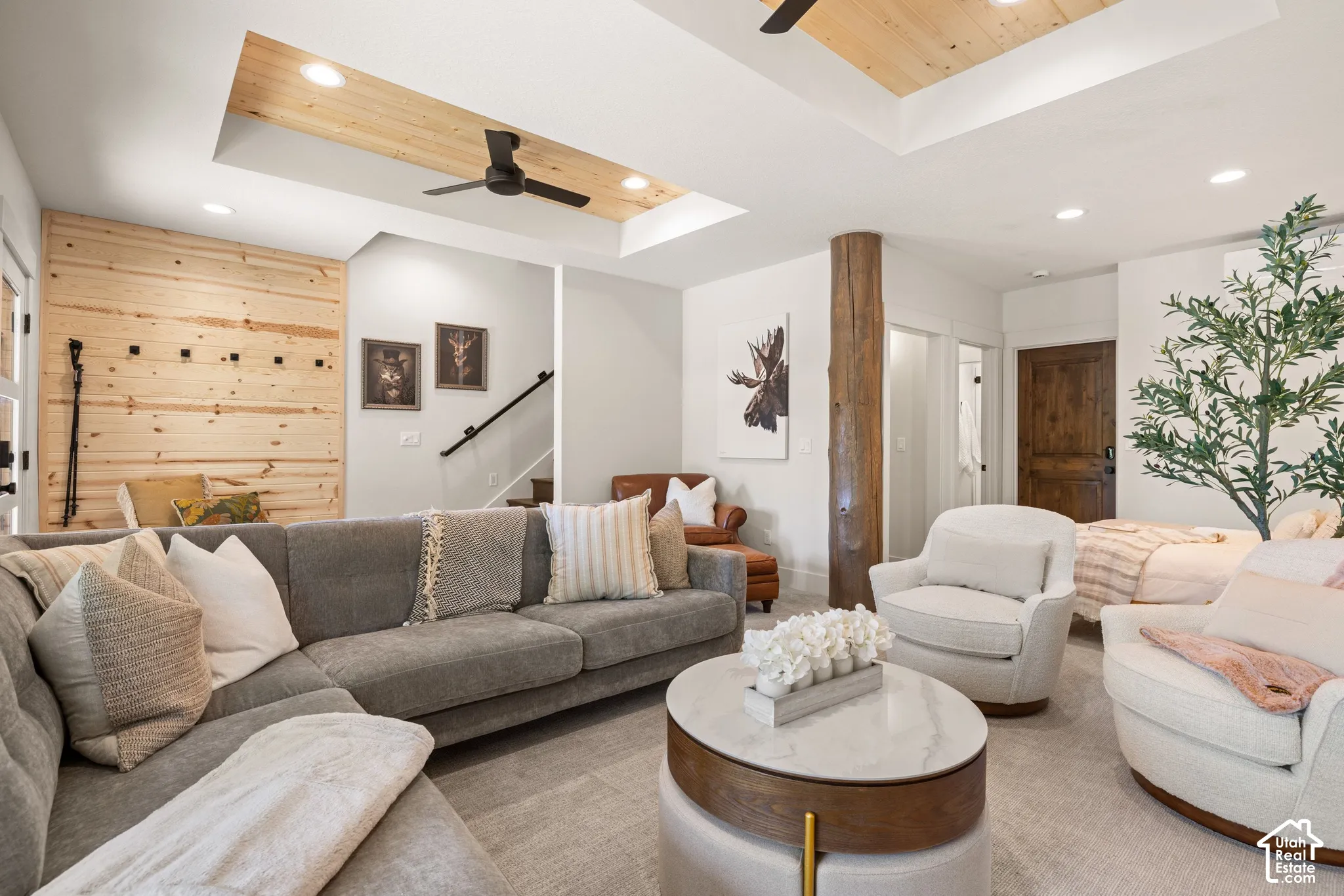 Recessed ceiling with wood-finish inlay and ceiling fan, wood-paneled accent wall, natural wood column, wood-finish interior door, and carpet flooring