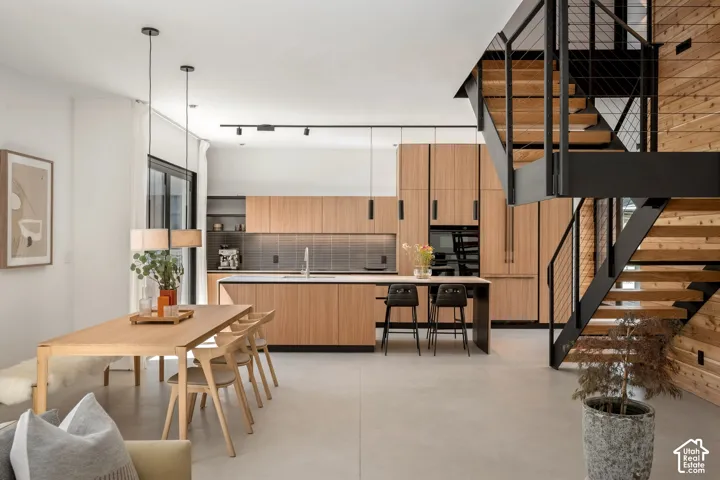 Open-concept living space with light wood-finish cabinetry, a large kitchen island, dark tile backsplash, and a contemporary open-riser staircase with metal railings