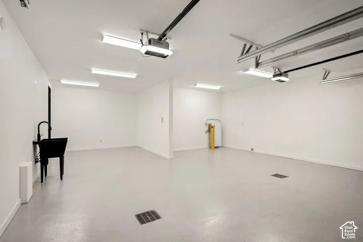 Spacious garage with light grey flooring, multiple overhead fluorescent light fixtures, a utility sink with a matte black faucet, and an automatic garage door opener system