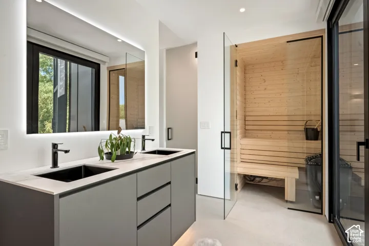 Double vanity with matte black fixtures, an illuminated mirror, and a built-in sauna with light wood paneling and a glass door