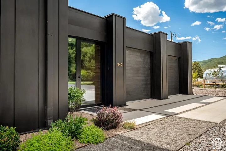Modern exterior featuring dark vertical siding, a full-light entry door, two garage bays with horizontal wood-finish doors, and a concrete paver driveway