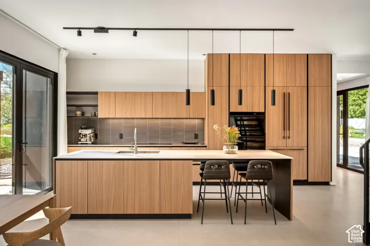 Modern kitchen featuring light wood-finish cabinetry, a large kitchen island with counter seating, integrated stainless steel appliances, and sleek black pendant lighting