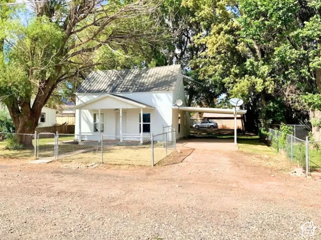 White exterior with a gable roof, covered carport, chain-link fencing, and mature trees surrounding the property