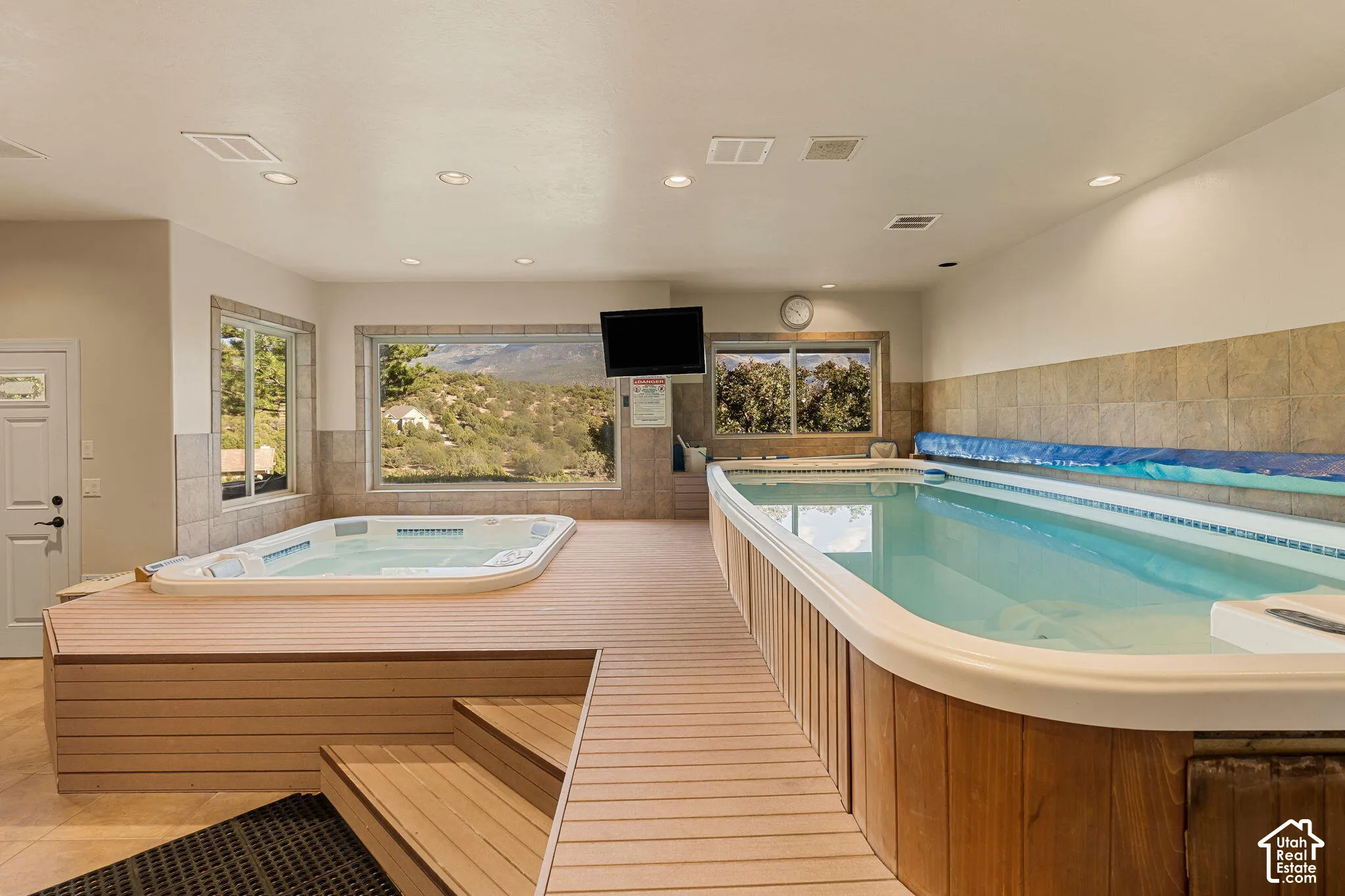 Indoor lap pool with integrated hot tub, surrounded by a light wood-finish deck with steps