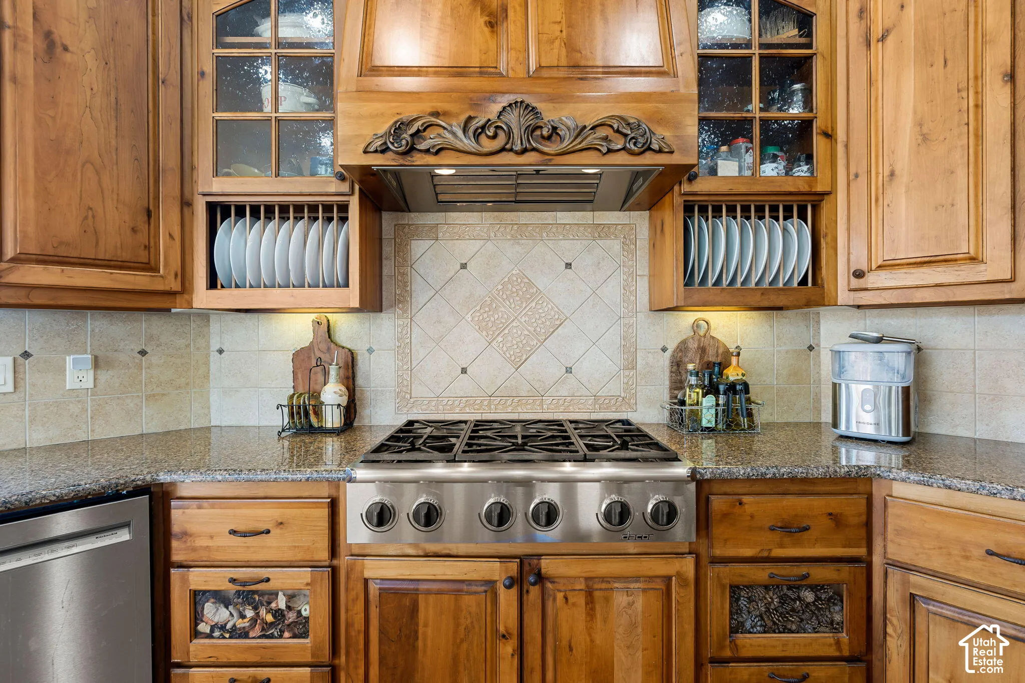 Wood cabinetry with glass-front display cabinets, stainless steel range with griddle, tile backsplash with decorative inset, and speckled countertops