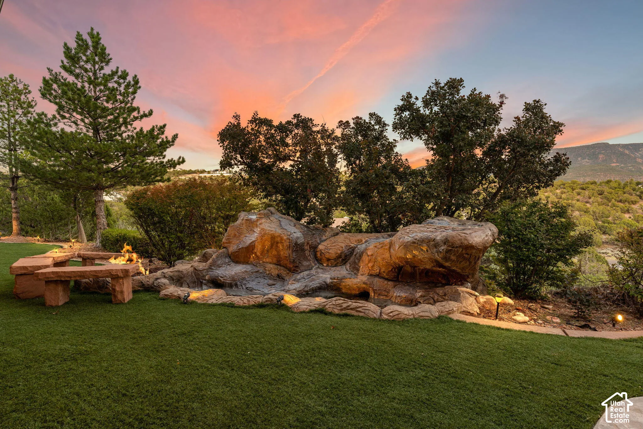 Expansive artificial turf lawn featuring a stone fire pit, built-in stone benches, and a rock waterfall with integrated lighting