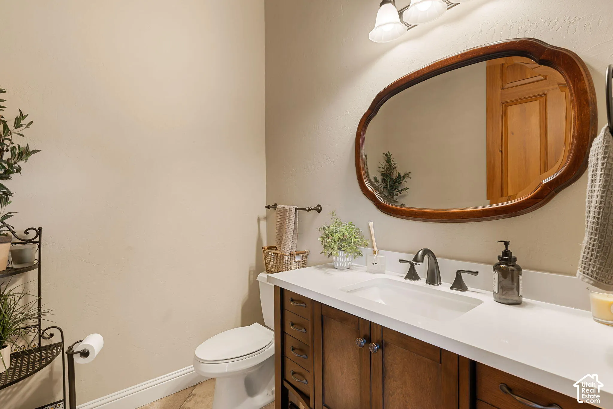 Wood-finish vanity with paneling and dark hardware, white countertop with integrated sink, dark bronze-finish faucet, and a decorative wood-framed mirror