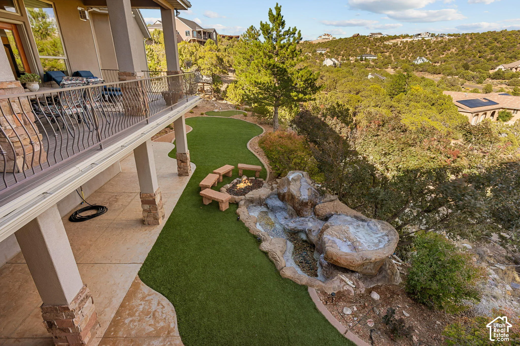 Expansive outdoor living area featuring a covered patio, wrought iron railing, artificial turf lawn, and a custom rock water feature