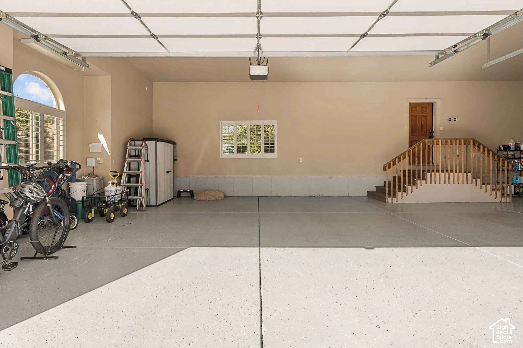 Expansive garage featuring a light-colored epoxy floor, an arched window with shutters, a rectangular window with shutters, interior access staircase, and a wooden entry door