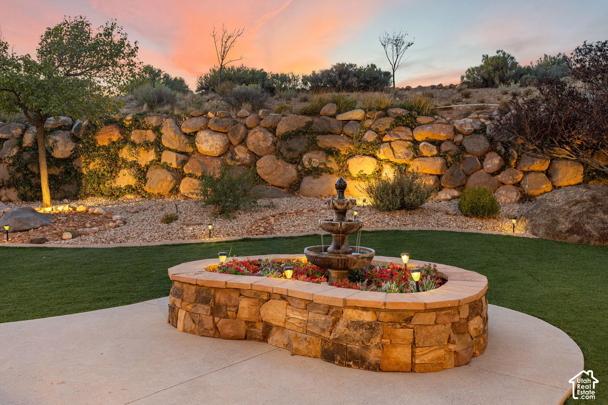 Tiered stone retaining wall with integrated lighting, artificial turf, concrete patio, and a stone-clad fountain planter