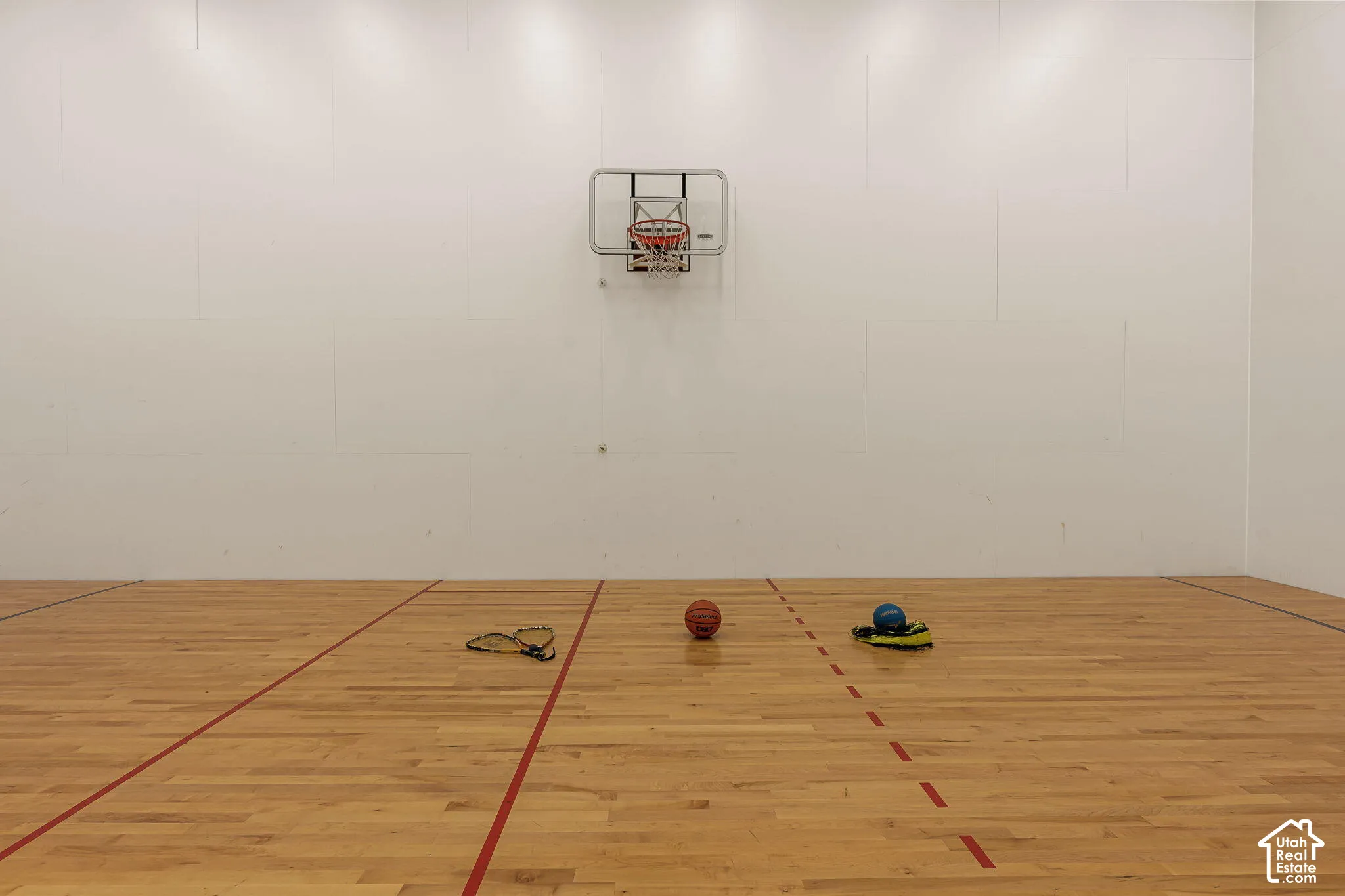 Indoor athletic court with light wood-finish flooring, white walls, and an attached basketball hoop