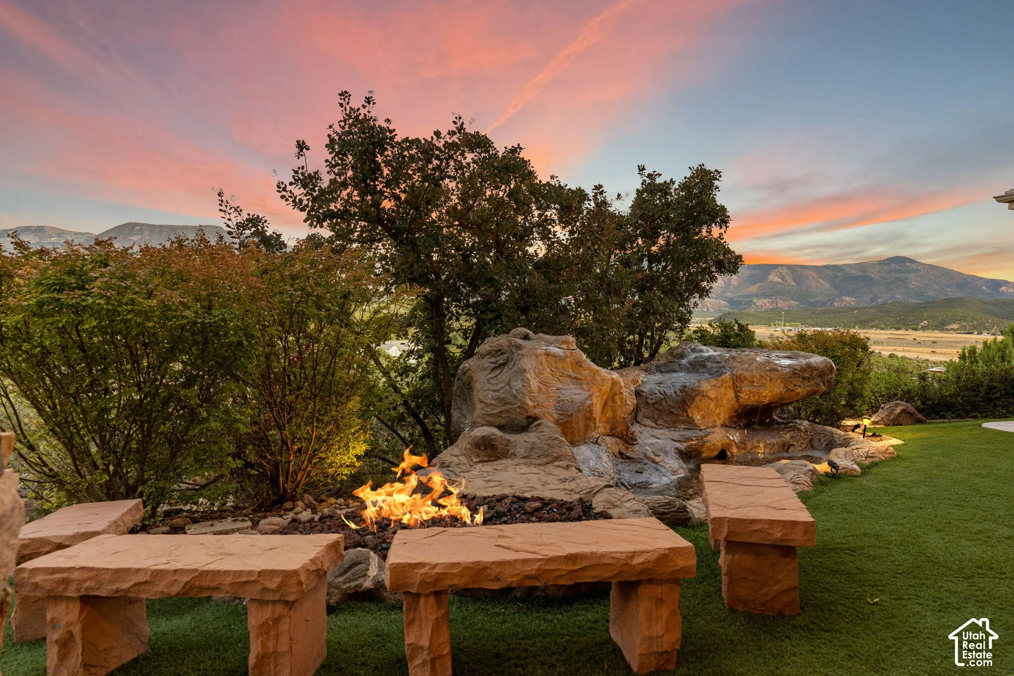 Built-in stone seating surrounding a fire pit, artificial turf, rock water feature, and mature landscaping