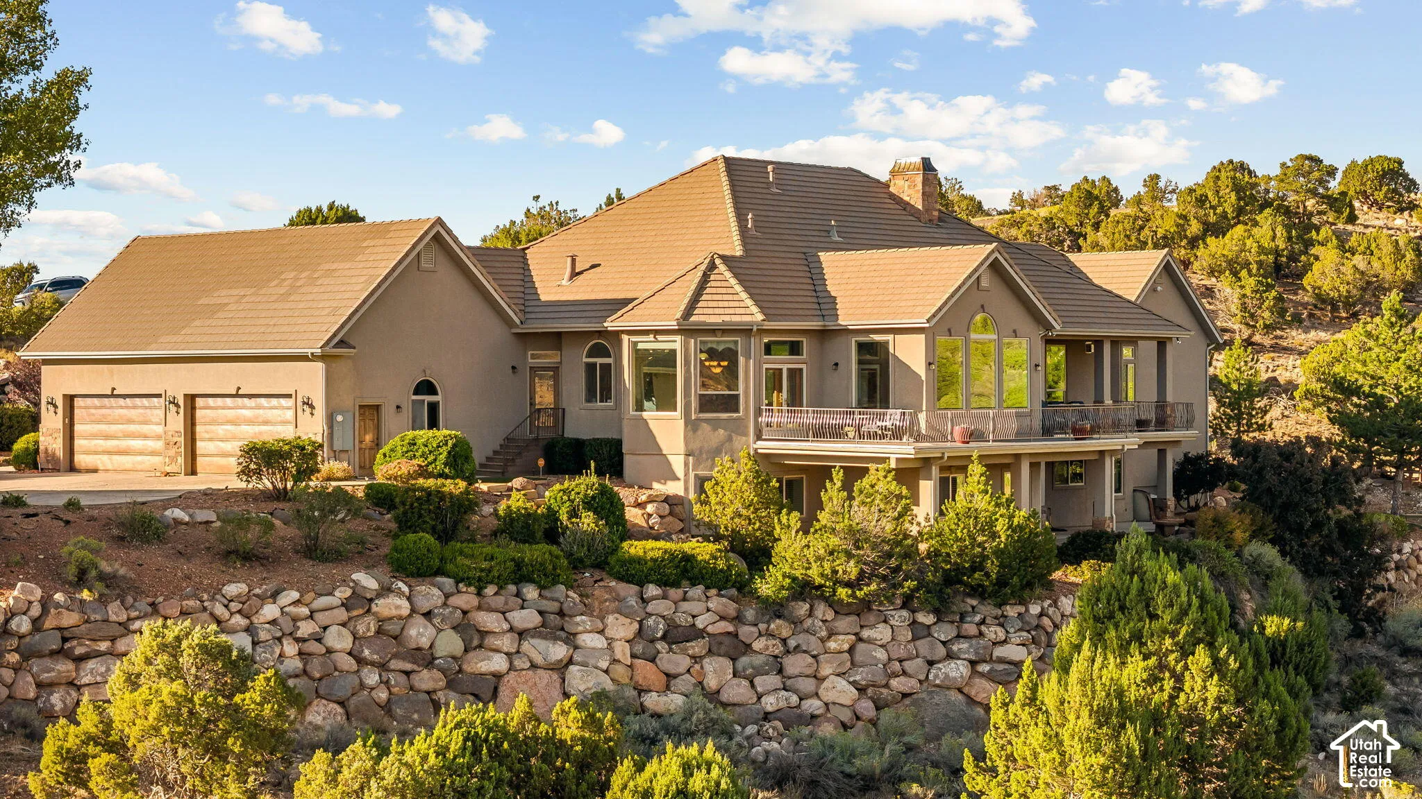 Stucco exterior with a tiled roof, multi-level balconies, an integrated two-car garage with wood-finish doors, and extensive stone retaining walls