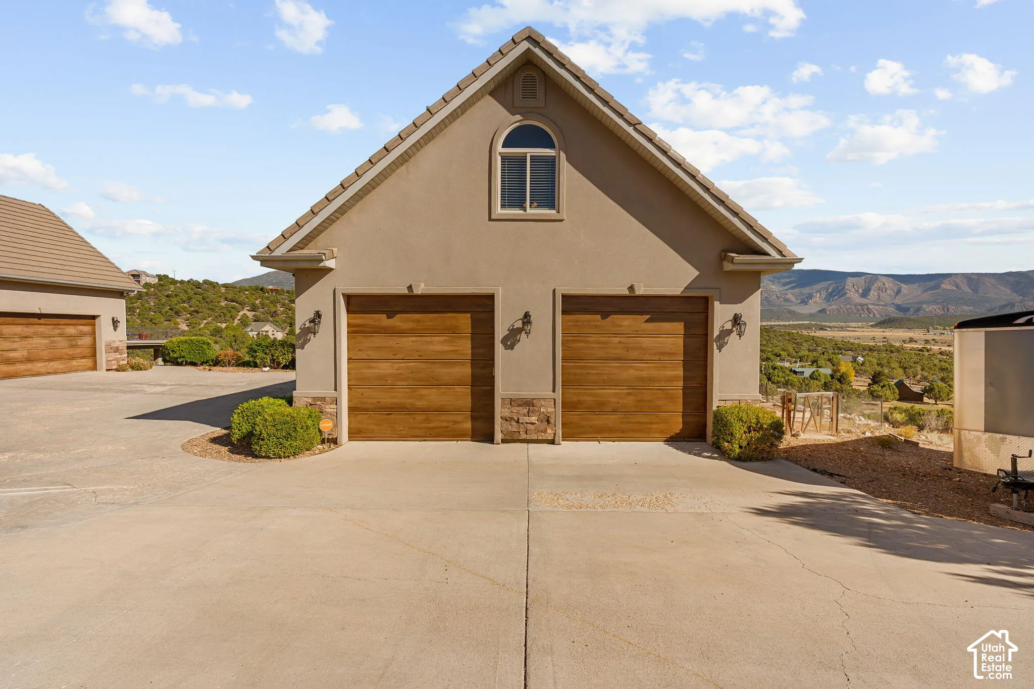 Two-car garage with wood-finish doors and stone accents, featuring an arched window and exterior sconces, situated on a paved driveway with distant mountain scenery