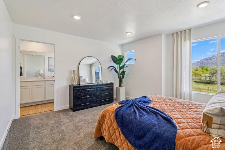 Recessed ceiling lighting, carpeted flooring, large window with mountain scenery, and an open doorway to an attached bathroom featuring a vanity with a white countertop