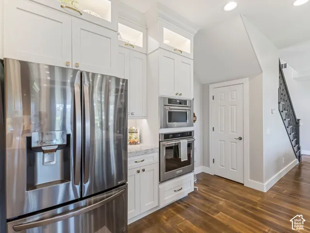 White cabinetry with glass display uppers, stainless steel French door refrigerator, built-in wall oven and microwave, wood-finish flooring, and a staircase with wrought iron balusters