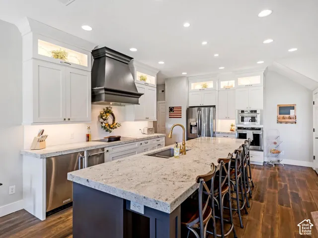 Expansive kitchen island with a stone-finish countertop, white cabinetry with upper display lighting, stainless steel appliances, and wood-finish flooring