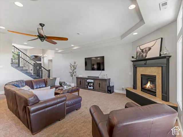 Fireplace with a stone surround and dark mantel, recessed lighting, wood-bladed ceiling fan, tray ceiling, and a staircase with a dark railing