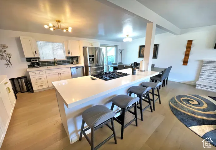 White shaker cabinetry, stainless steel appliances, light wood-finish flooring, a white countertop island with seating, and a geometric chandelier