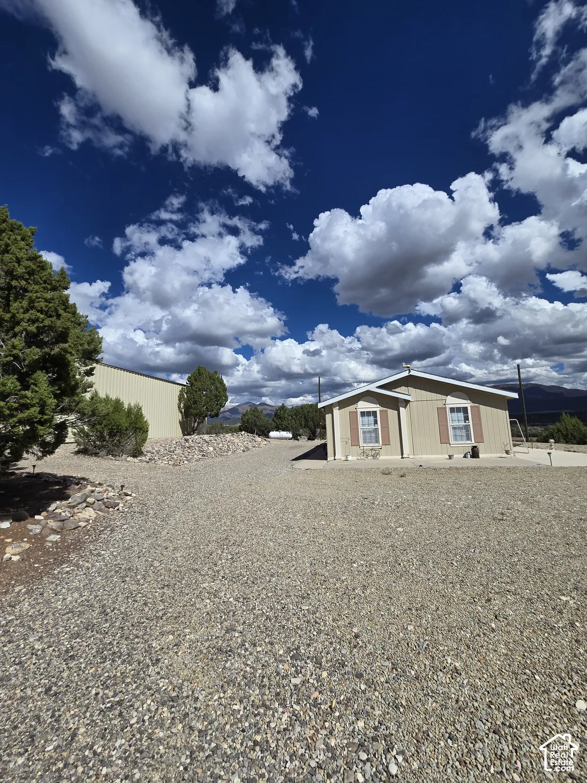 Gravel driveway with a single-story structure featuring tan siding and brown window shutters