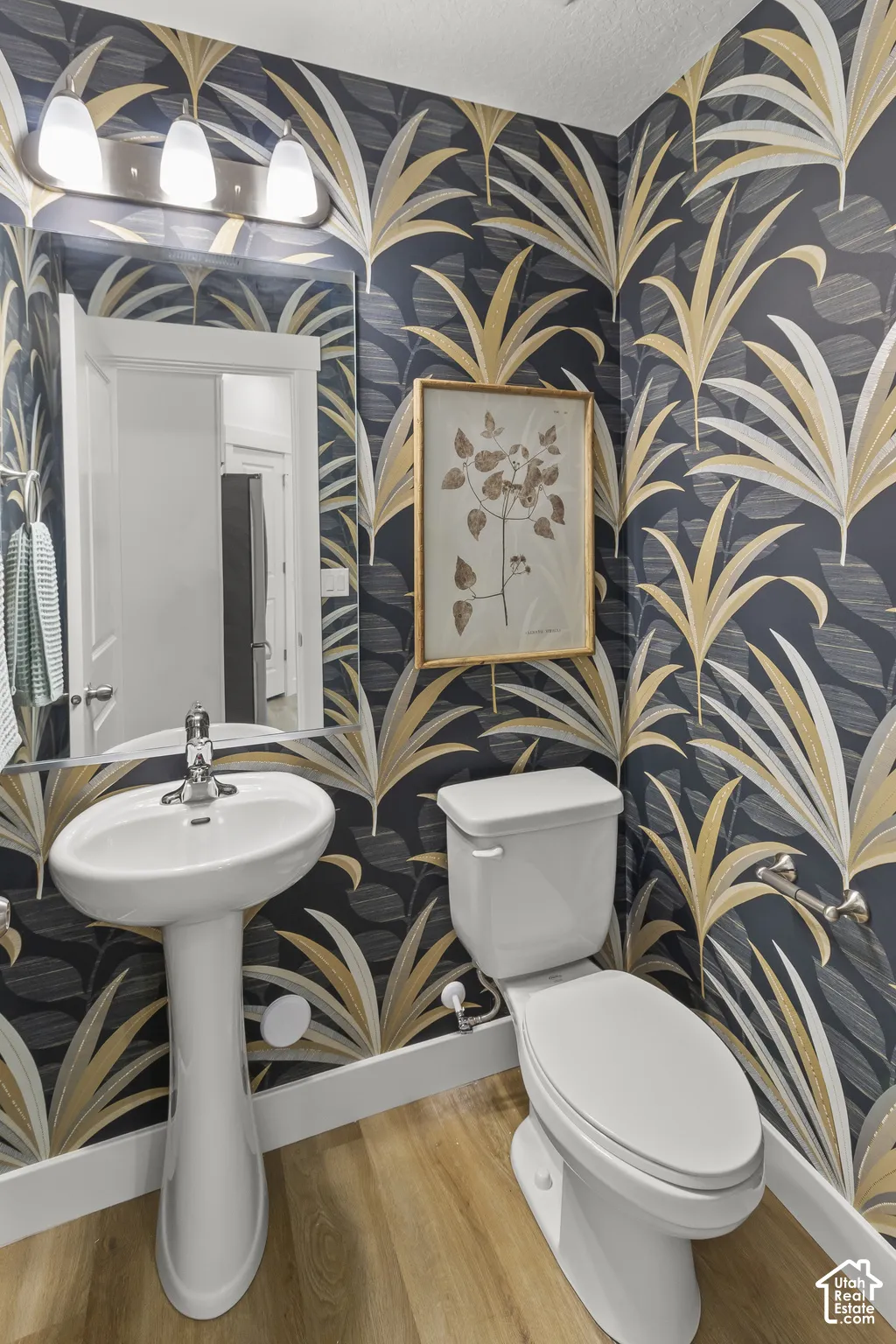 Pedestal sink, wall-mounted mirror, overhead vanity lighting, wood-finish flooring, and a contemporary wallpaper design