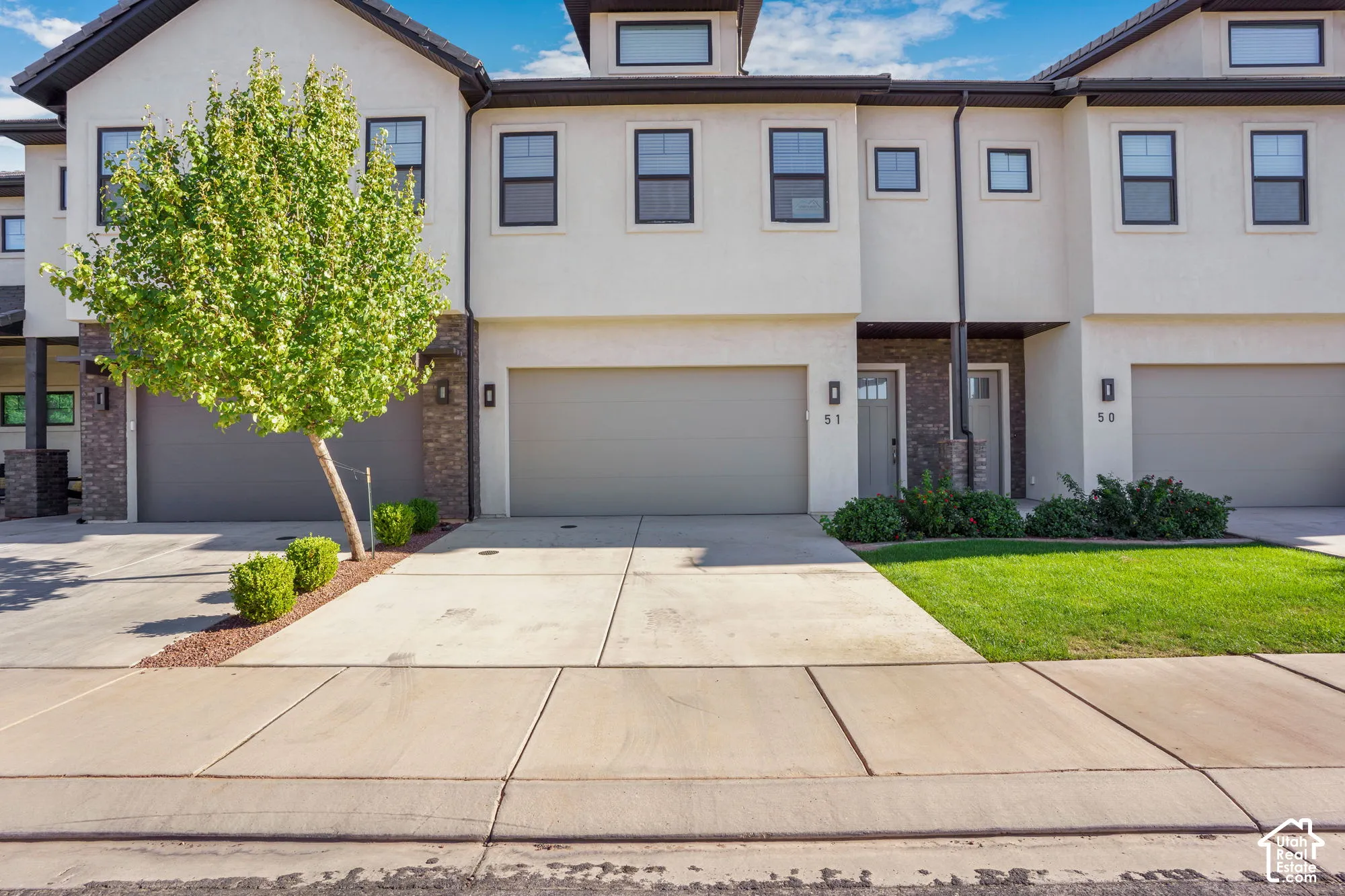 Stucco exterior with dark trim accents, integrated single-car garages, mature landscaping with a tree and shrubs, and concrete driveways